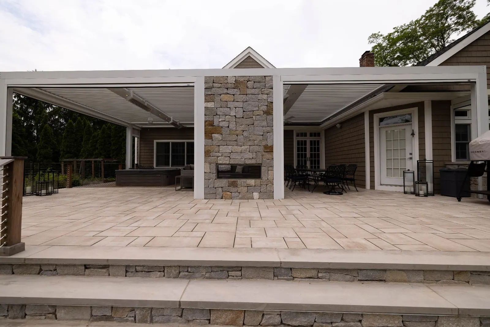 White louvered pergola spanning a patio with a central stone fireplace