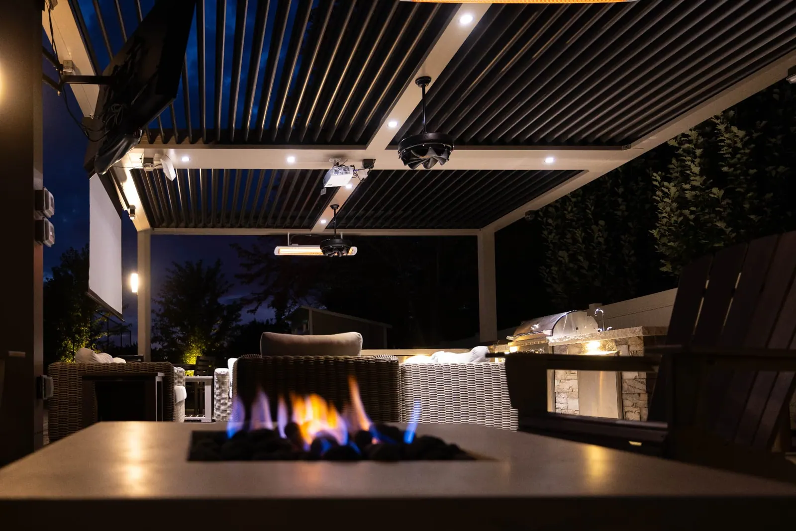 Illuminated louvered pergola above an outdoor kitchen and fire table at night
